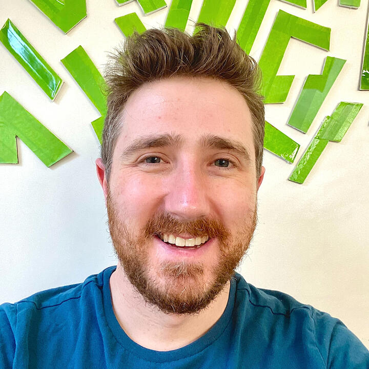 RJ Foley with a relaxed smile on his face. His beard and hair are a mix of slightly dirty blonde and golden auburn, styled naturally with a flip at the front of his scalp which complements his teal shirt. Behind him are some shapes sticking up on the wall